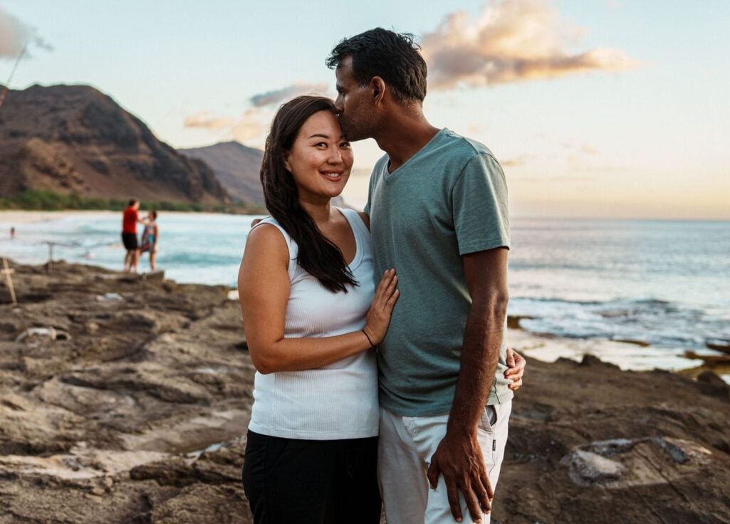 A man kisses a smiling East Asian woman on the forehead as they embrace on a rocky coastline at sunset, evoking the serene and beautiful landscapes people seek out when researching the best places to retire in the world.