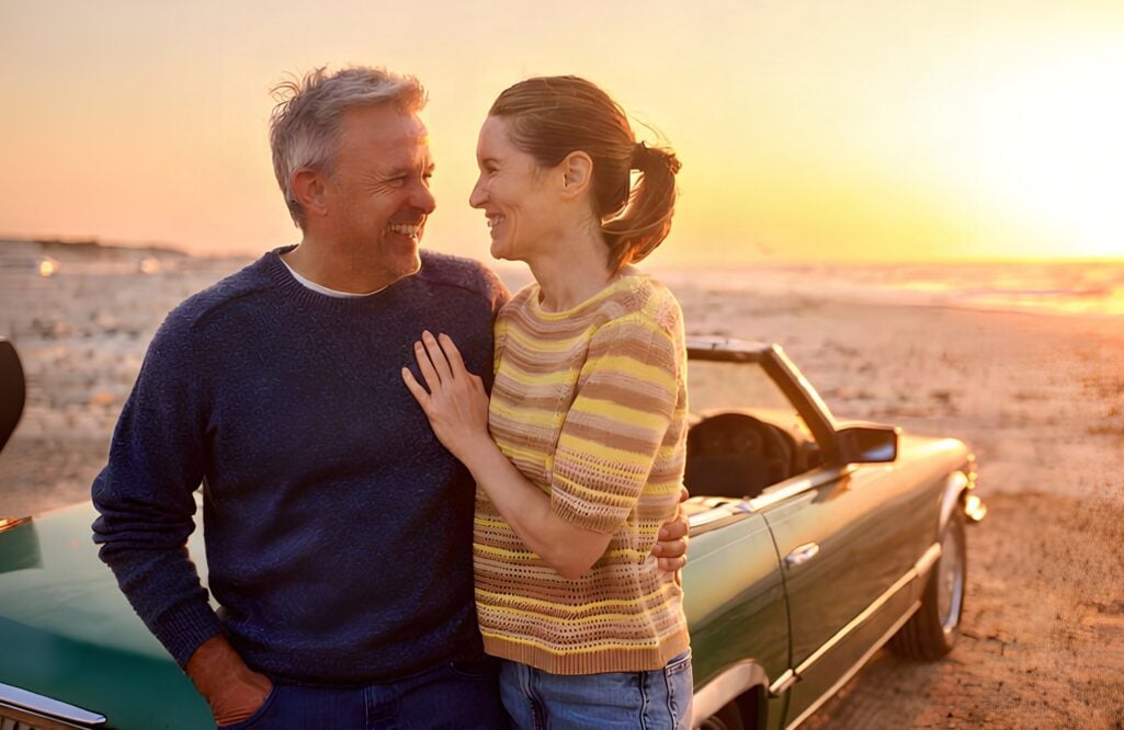 A smiling couple embraces next to a convertible car on a sandy beach at sunset, enjoying the freedom and adventure of life after retirement.