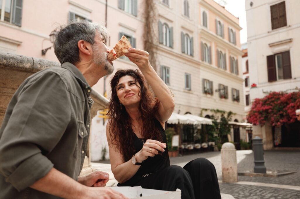 A mature woman feeds a slice of pizza to a man while sitting by a fountain in a European city plaza, capturing the vibrant lifestyle and culture enjoyed by those who choose to retire abroad.