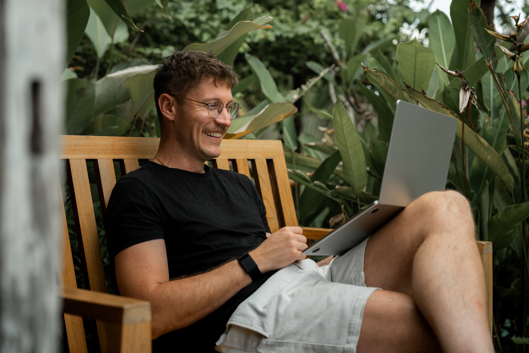 A smiling man with glasses sits on a wooden bench outdoors with his laptop, enjoying a relaxed moment while testing his money management knowledge with a financial literacy quiz.