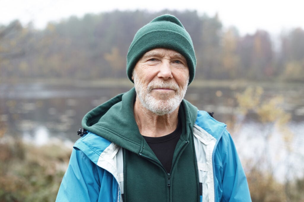 A senior man with a thoughtful expression stands by a tranquil lake in autumn, wearing a warm beanie and jacket. His calm yet serious gaze reflects the resilience needed when navigating financial uncertainties like potential social security benefit cuts.