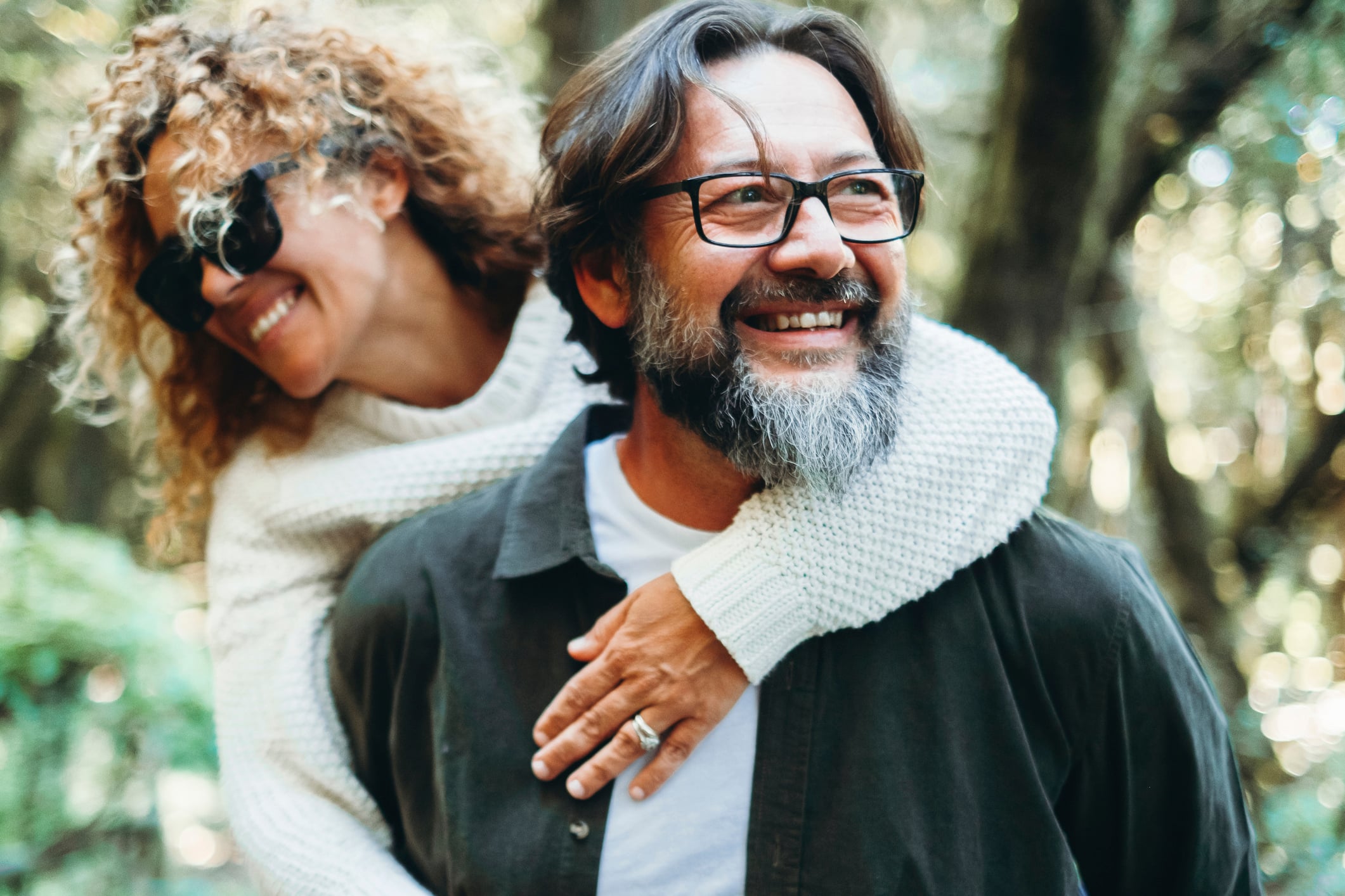 A happy, smiling middle-aged couple enjoying a piggyback ride outdoors in a sunlit wooded area, representing the freedom and joy that come when you are able to retire with confidence.