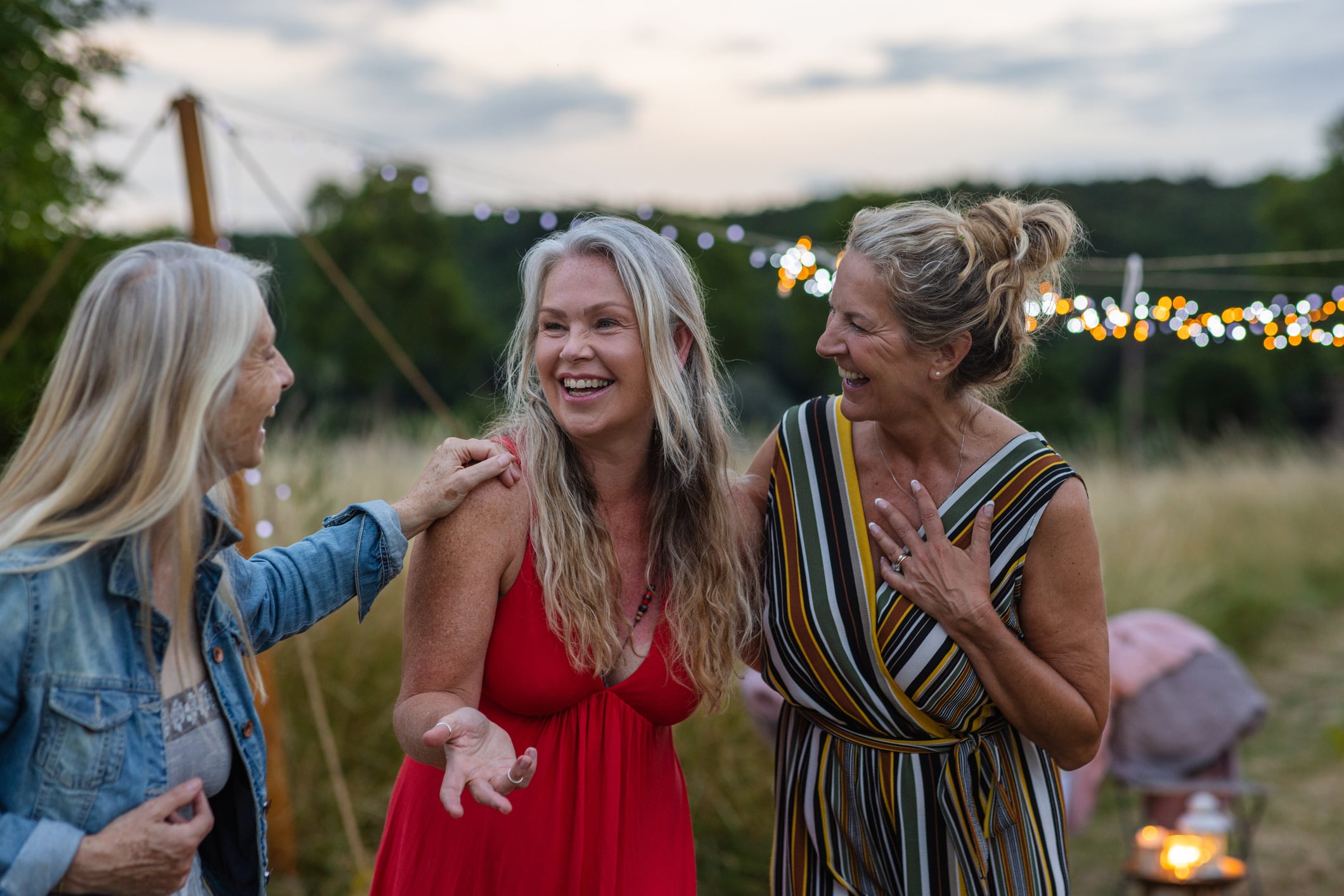 Three joyful, elegantly dressed women with silver hair laugh together at an outdoor evening garden party, representing the vibrant social lives and financial freedom often enjoyed by the wealthiest retirees.