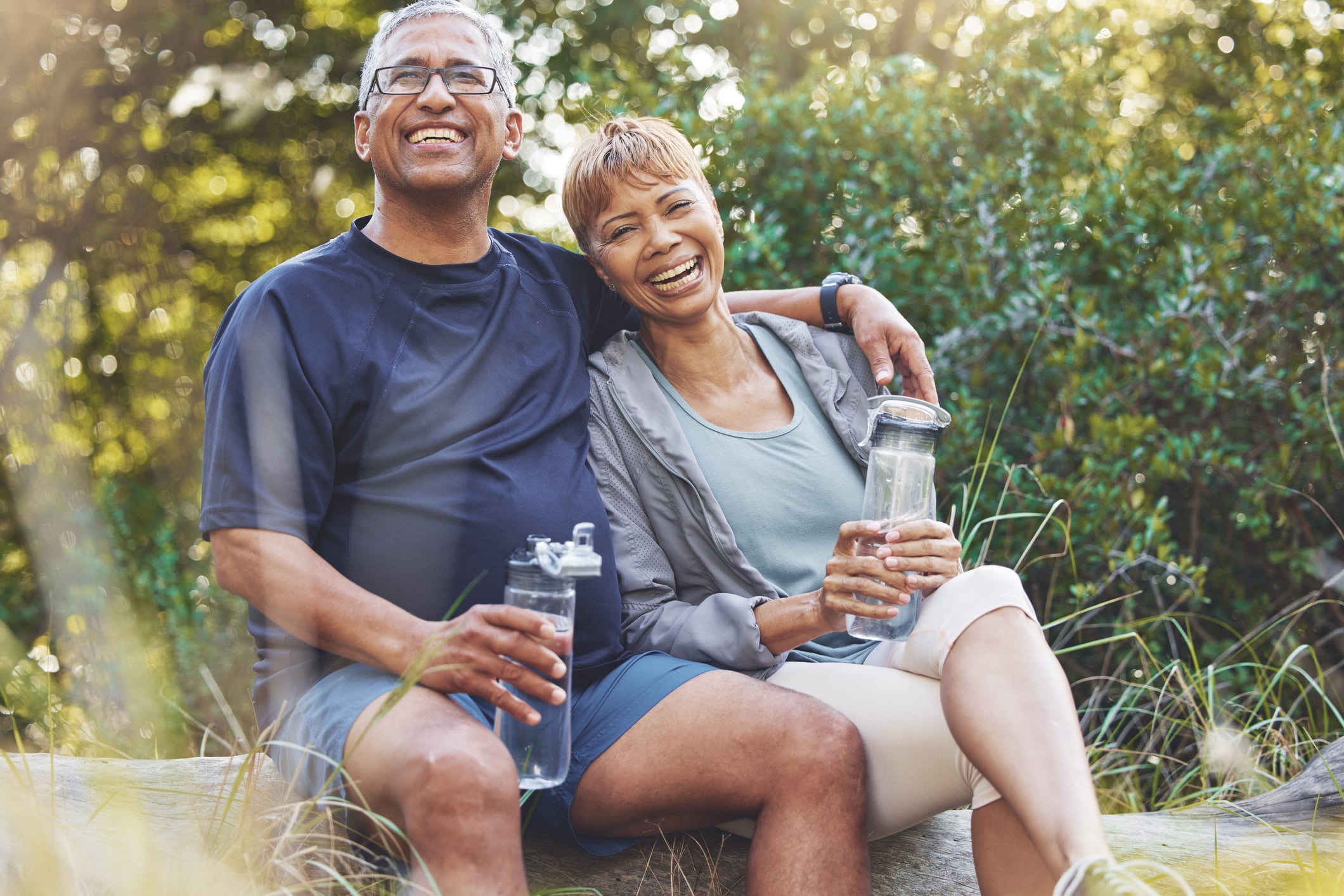 A happy senior couple sits together on a log laughing while holding water bottles during a hike, illustrating how reliable retirement income can help bridge the gap left by a low S&P 500 dividend yield.