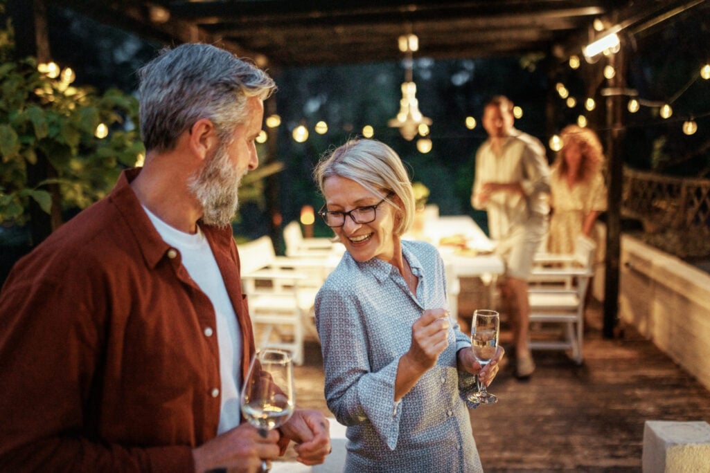 An older couple smiles and chats while holding wine glasses during an evening patio dinner party, with a younger couple in the background, capturing the joy of celebrating major family milestones like buying a house for your child.