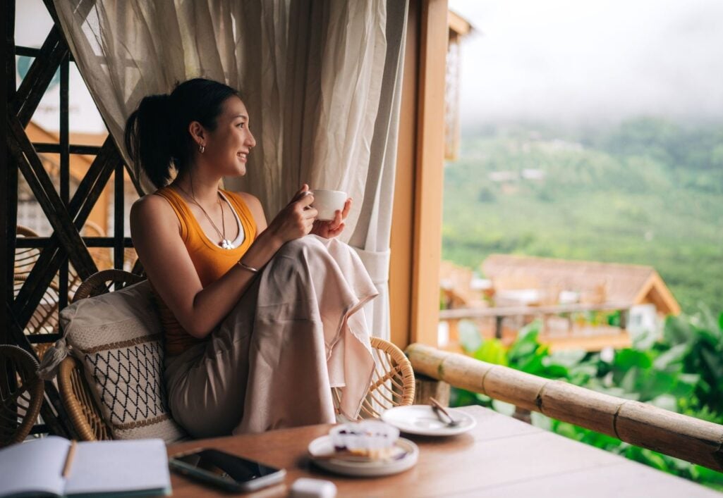 A smiling woman rests on a wicker chair with a mug on a balcony overlooking lush, foggy mountains, capturing a quiet moment of personal freedom and how how wealth is defined beyond just money.