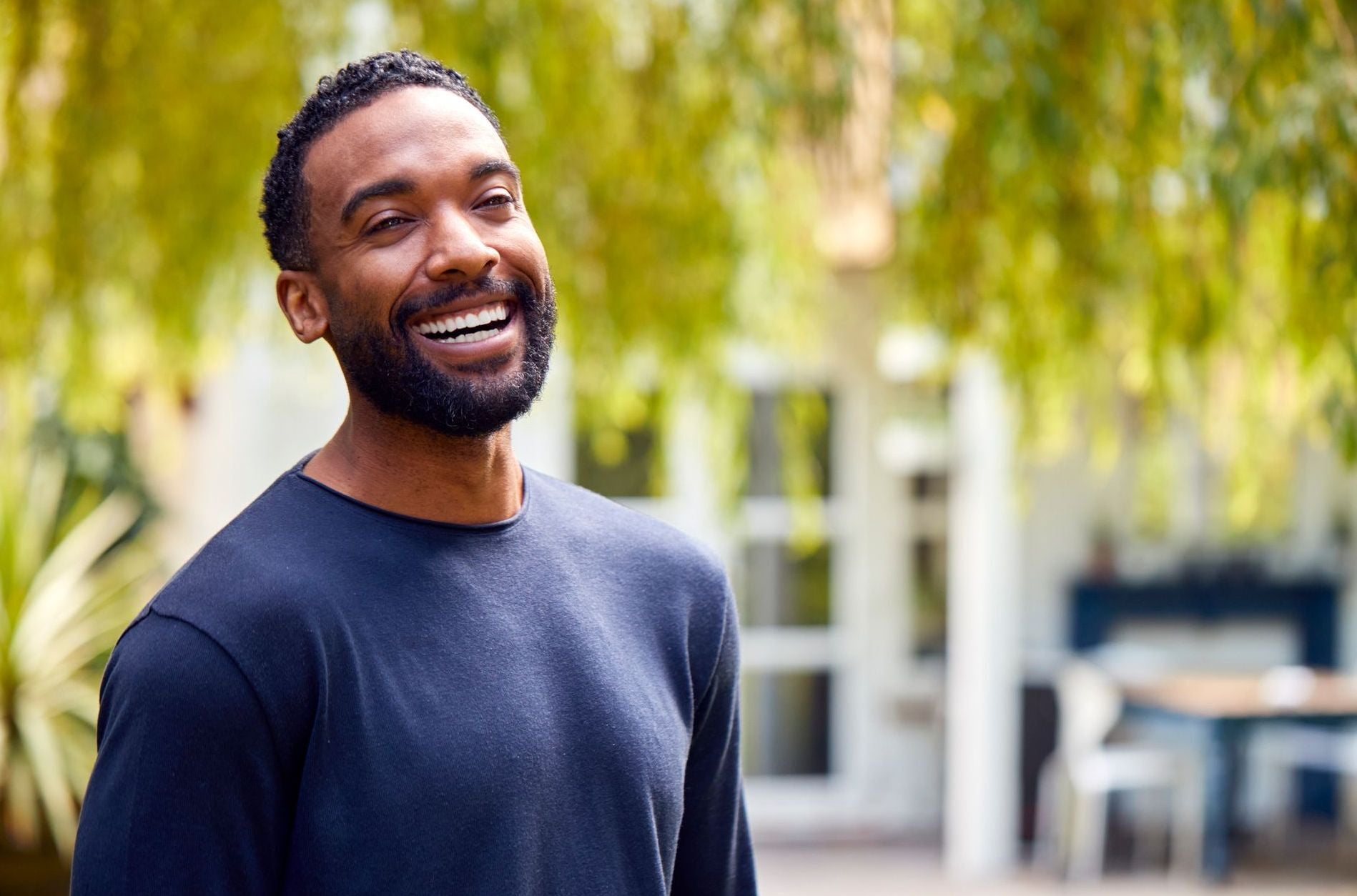 A smiling Black man with a beard looks off-camera while standing outside in a sunlit garden, reflecting the joy and freedom of planning early to beat the average retirement age.