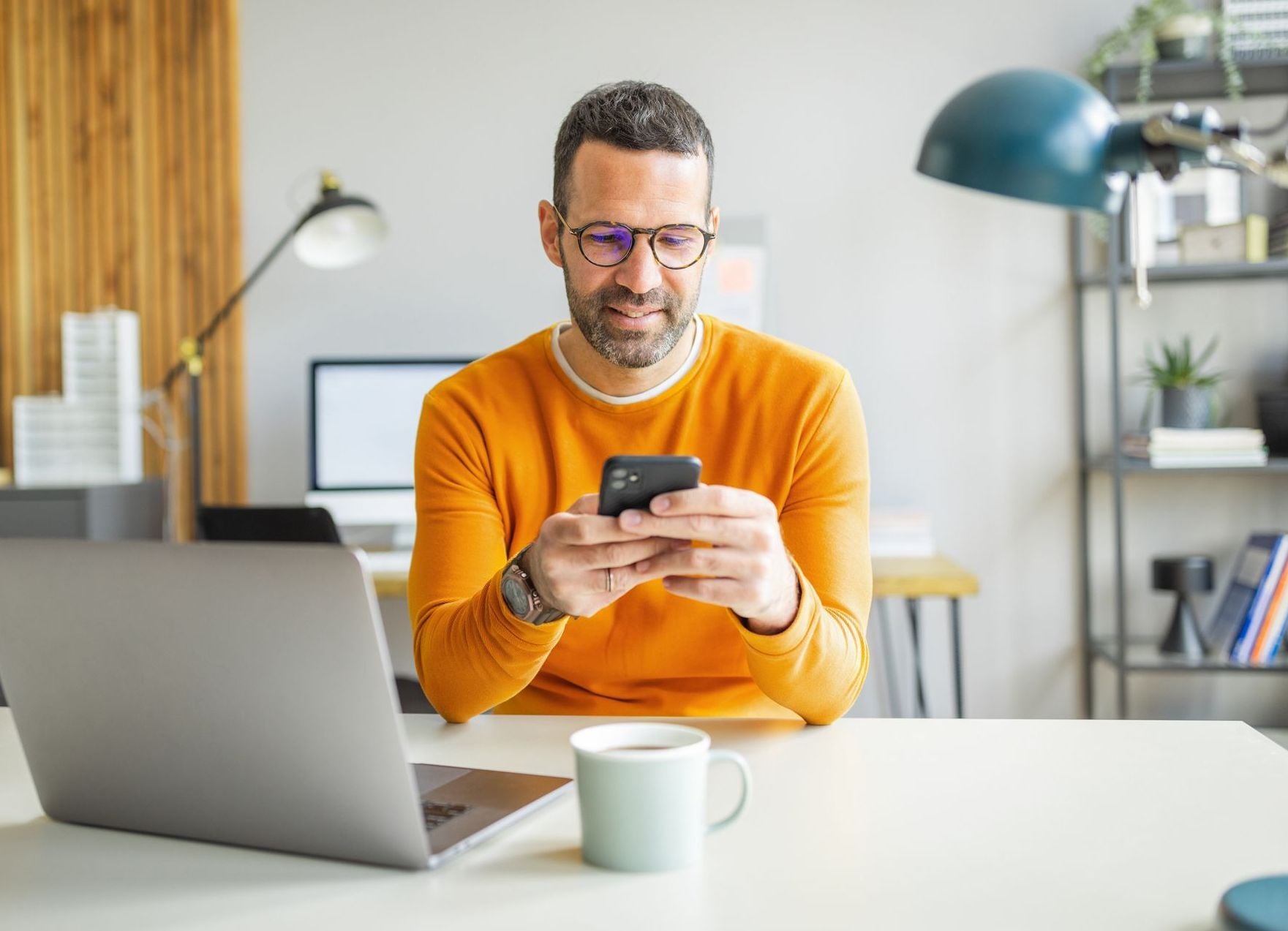 A man in an orange shirt smiles while looking at his smartphone at his desk with a laptop, comparing digital financial tools like Boldin vs. Empower to optimize his retirement portfolio.