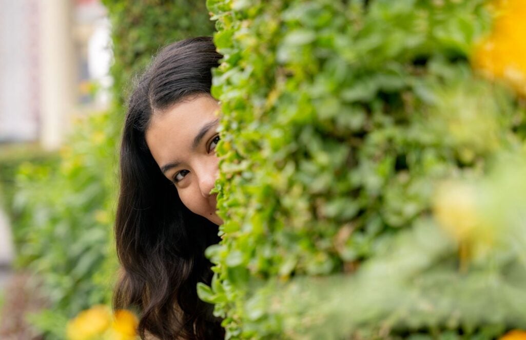 A young woman playfully peeks out from behind a lush green hedge, representing how uncovering hidden financial strategies can lead to greater long-term financial well-being.