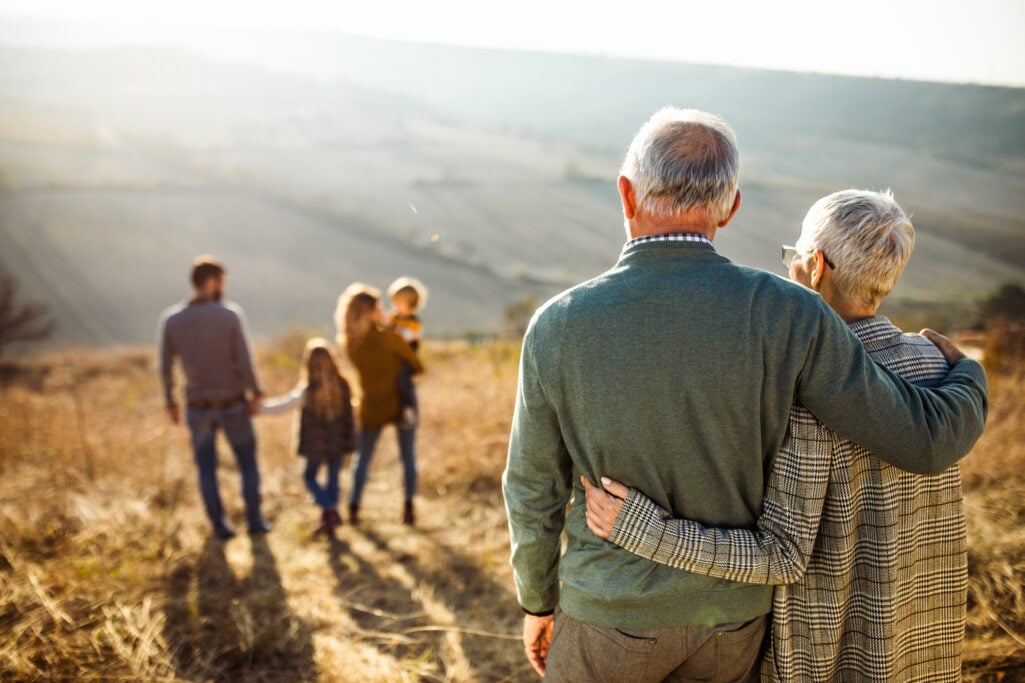 An older couple stands arm-in-arm from behind, looking out over a rolling countryside hill at their adult children and grandchildren, symbolizing the lasting family legacy and transfer of baby boomer wealth.
