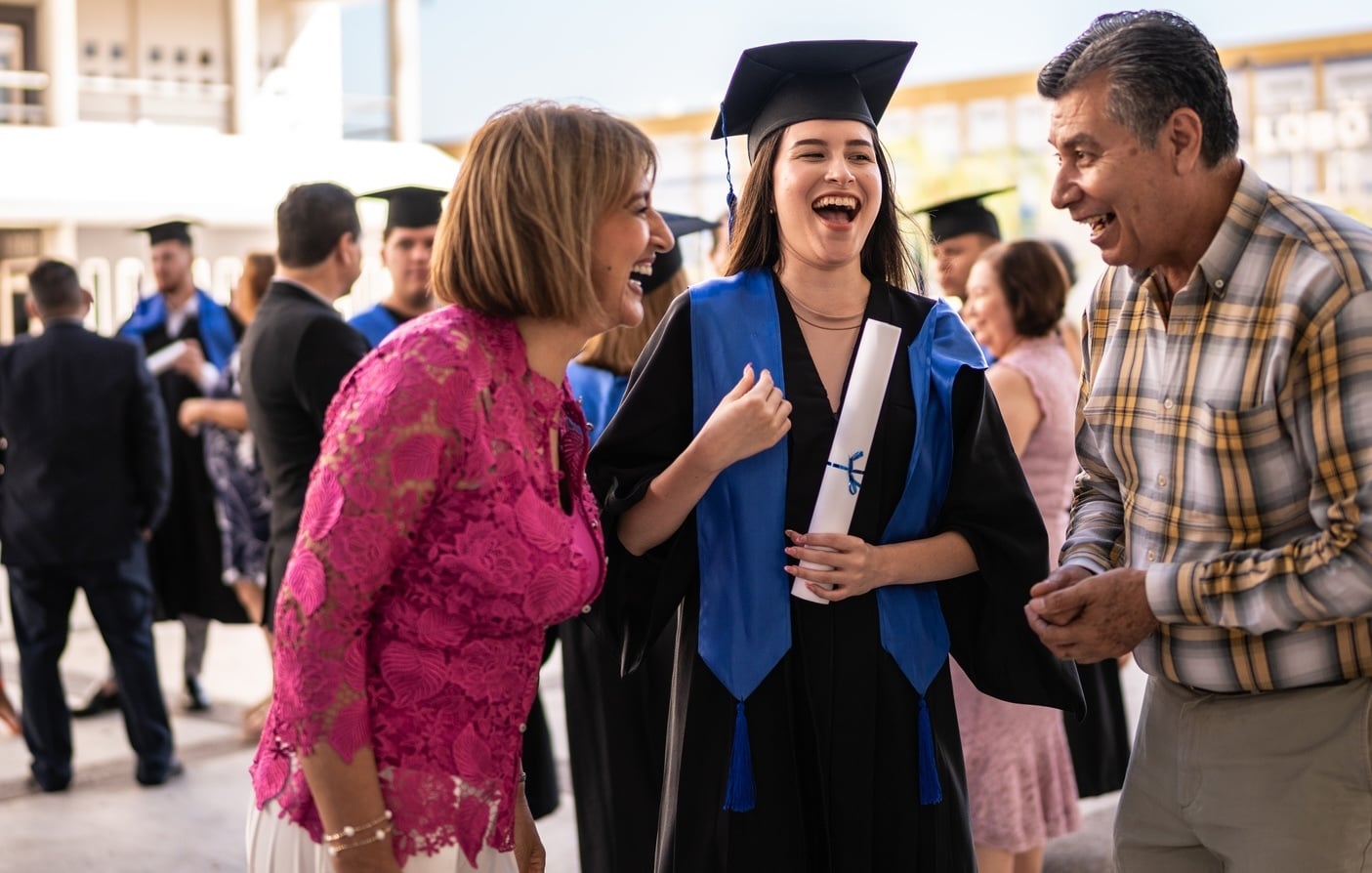 A young female graduate in a cap and gown laughs joyfully with her parents outdoors at her graduation ceremony, highlighting a major family milestone and prompting parents to consider what to do with money after kids graduate.