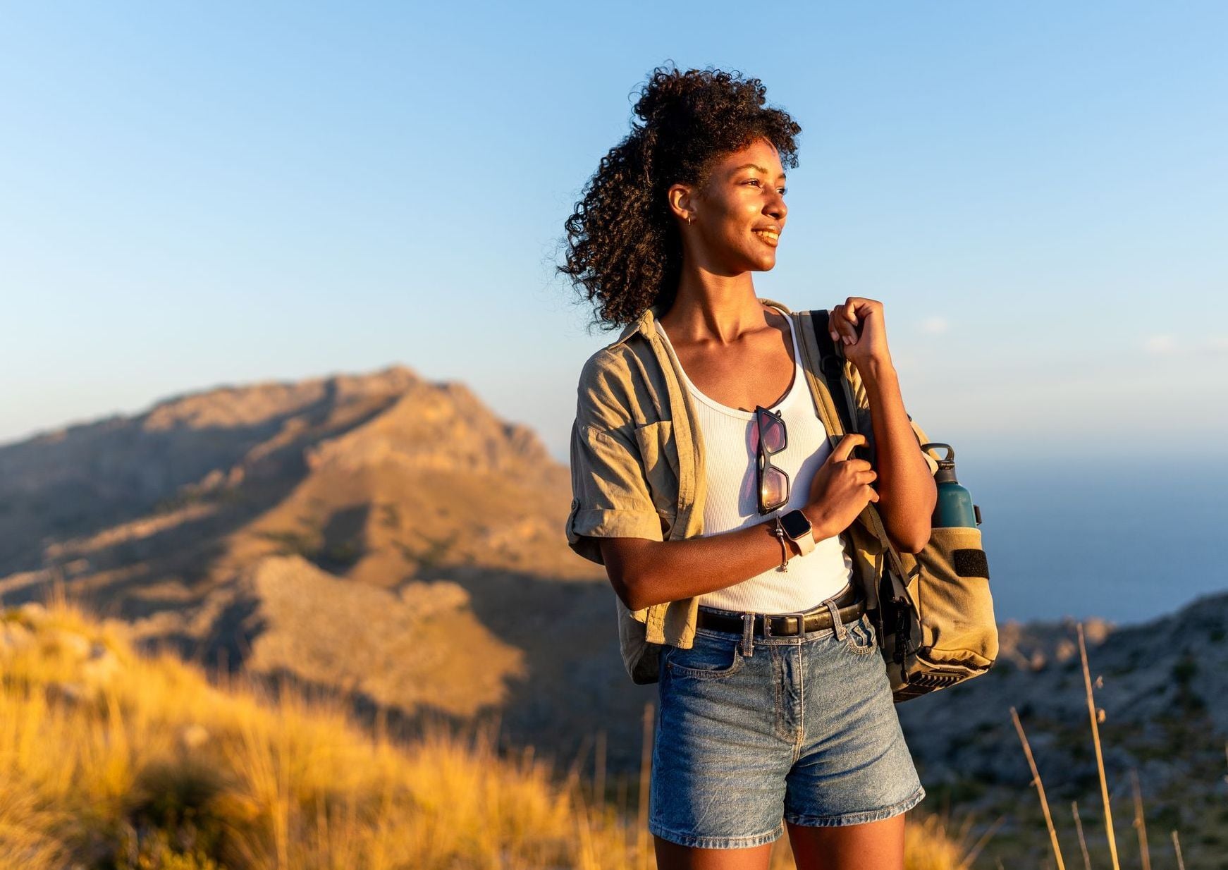 A smiling young Black woman with curly hair hikes along a grassy mountain ridge at sunset, carrying a backpack and looking toward the horizon, symbolizing a strong start toward building a healthy average 401k balance by age.