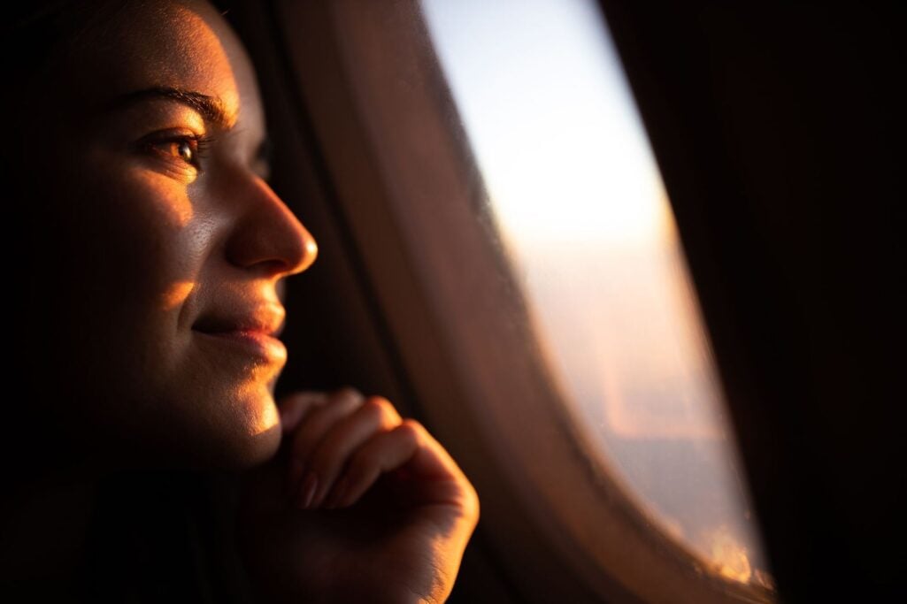 A close-up profile of a thoughtful woman looking out an airplane window at sunrise, capturing a moment of reflection on personal goals and the underlying money beliefs that shape financial journeys.
