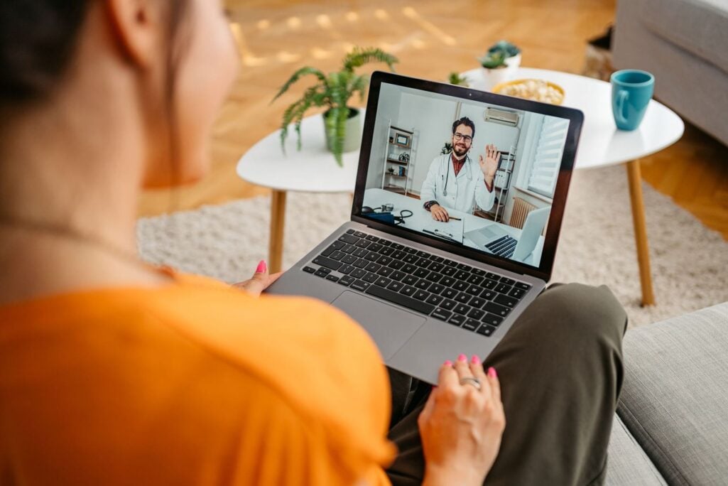 A woman sits on a sofa during a video appointment with a doctor on her laptop, representing the routine medical expenses that make up the average cost of healthcare in retirement.