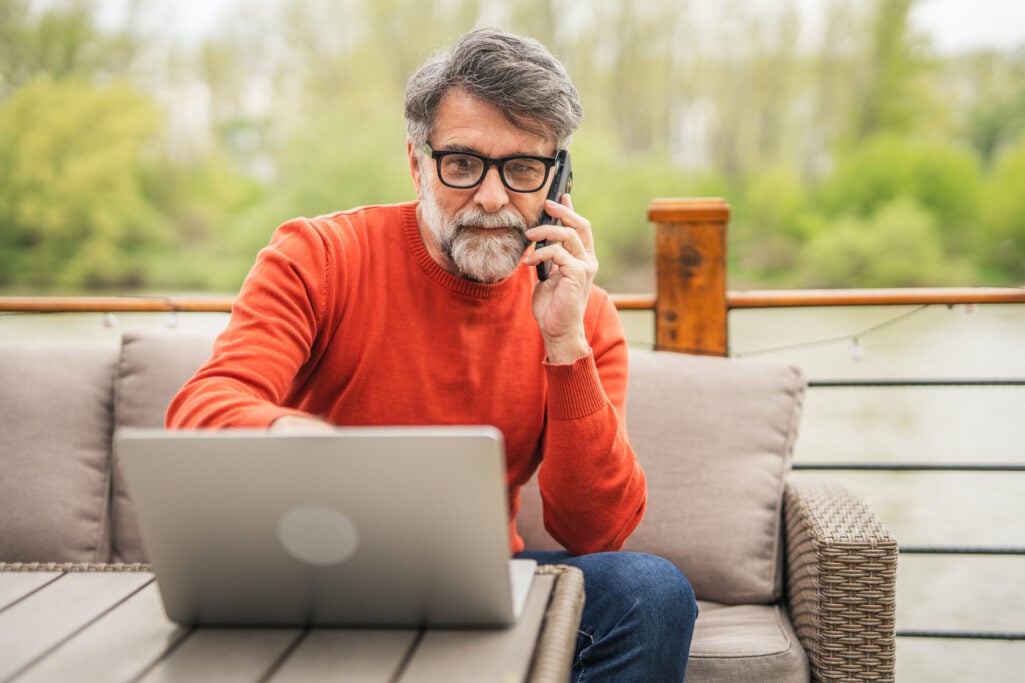 A middle-aged man with glasses speaks on the phone while working on his laptop on an outdoor patio, evaluating his knowledge before taking an online financial skills test.