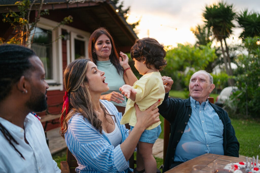 A multi-generational family smiles and gathers around a toddler at an outdoor birthday celebration, illustrating a milestone moment when parents or grandparents might evaluate whether a Trump Account is worth it.