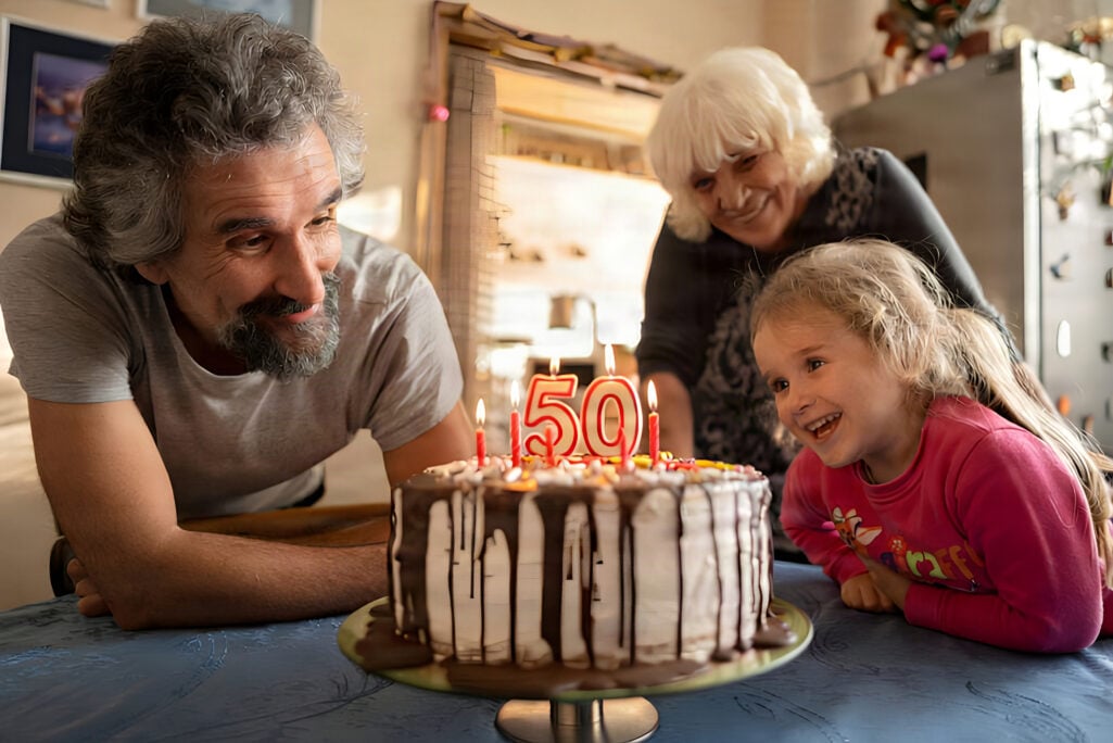 A family gathers around a 50th birthday cake as a man prepares to blow out the candles, marking a key milestone age for catch-up contributions when tracking retirement milestones by age.