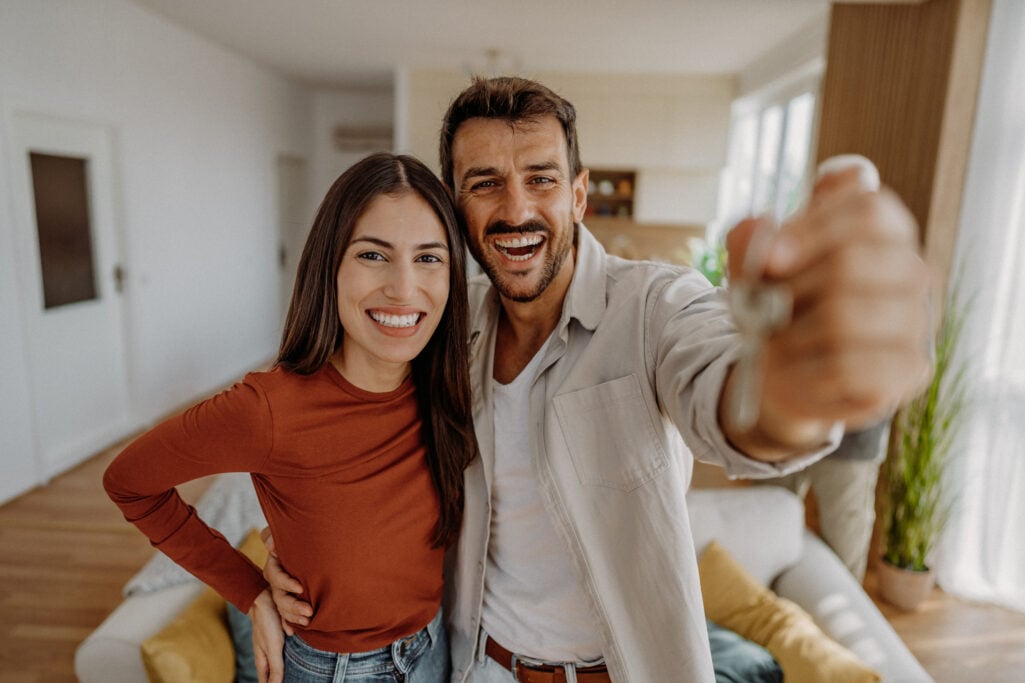 A cheerful couple holds up keys inside their house, celebrating homeownership as a primary path toward building higher than average home equity over time.